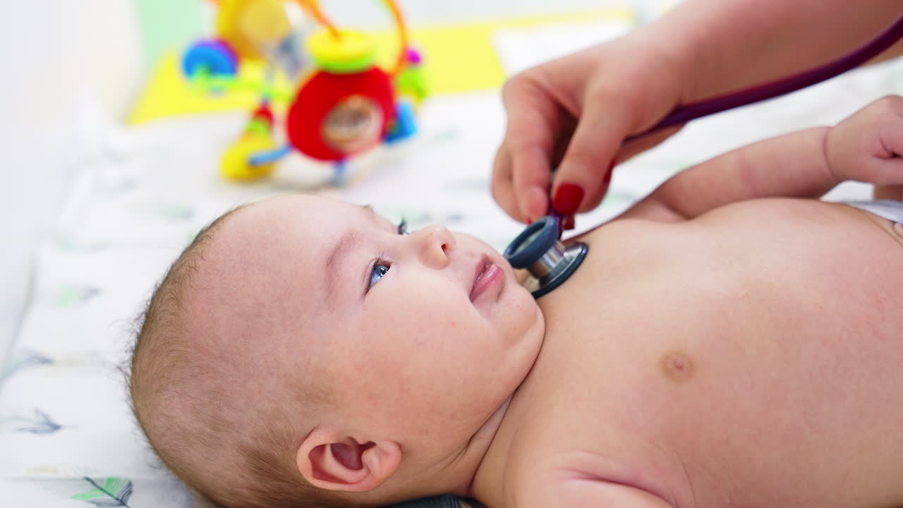 Pediatrician hand puts stethoscope on child's chest. Baby looks interested and smiles to the doctor. Blurred backdrop.