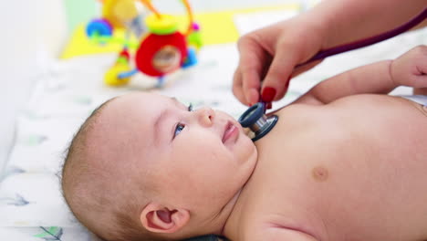 pediatrician hand puts stethoscope on child's chest. baby looks interested and smiles to the doctor. blurred backdrop.