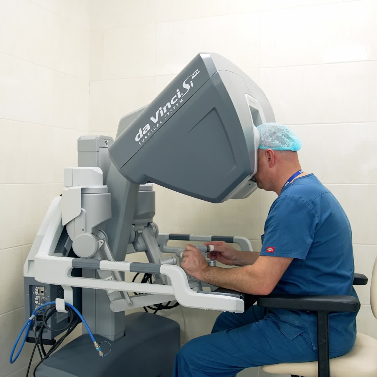 Medical professional in blue uniform and cap sitting at apparatus. Distancing from a doctor operating modern futuristic Da Vinci robot