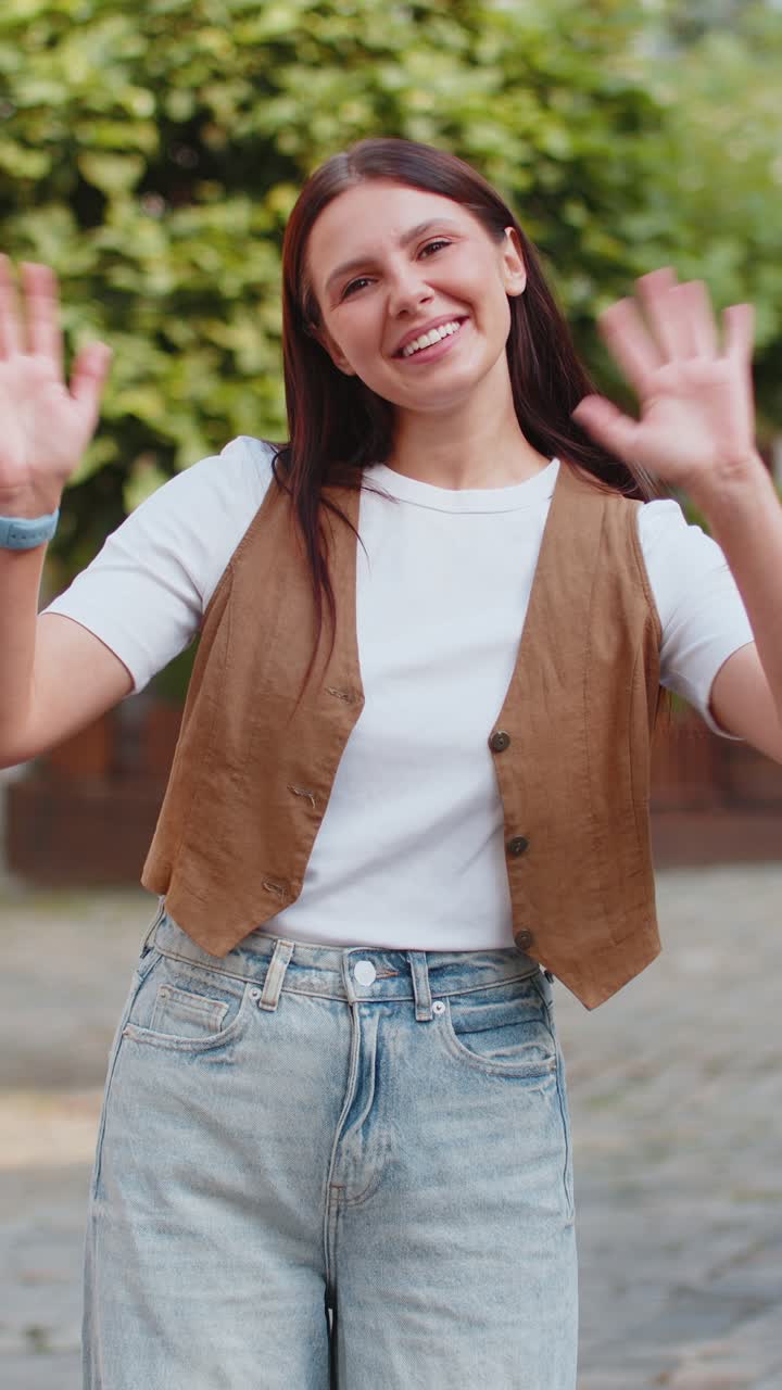 Cheerful young woman smiling friendly at camera waving hands gesturing while standing on city street