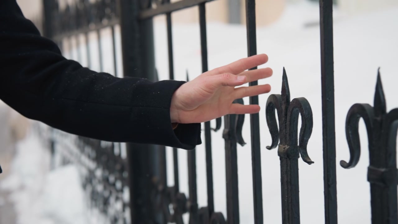 Nærbillede af en persons hånd, der rækker ud efter og rører ved et udsmykket sort metalhegn i snevejr. Scenen viser interaktionen mellem menneskelig berøring og en smedejernsport med sne synlig på jorden.