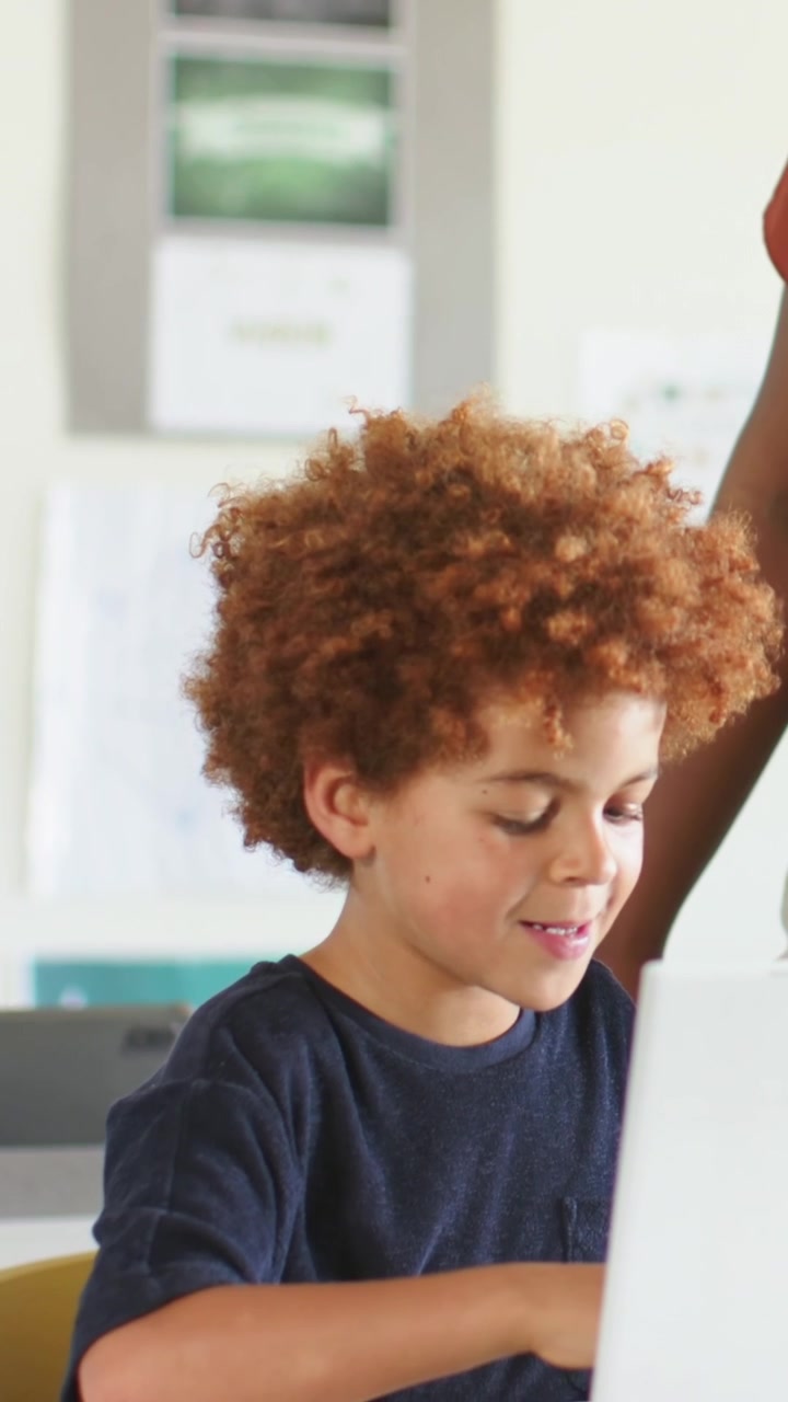 Vertical video: Focusing child in navy tee typing homework on silver laptop, glancing at rust adult