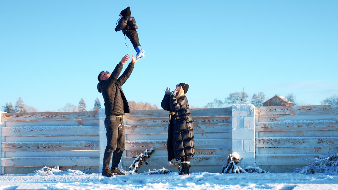 Happy Caucasian father tosses his baby boy outdoors in winter. Mother stands beside clapping hands. Family fun time in winter.