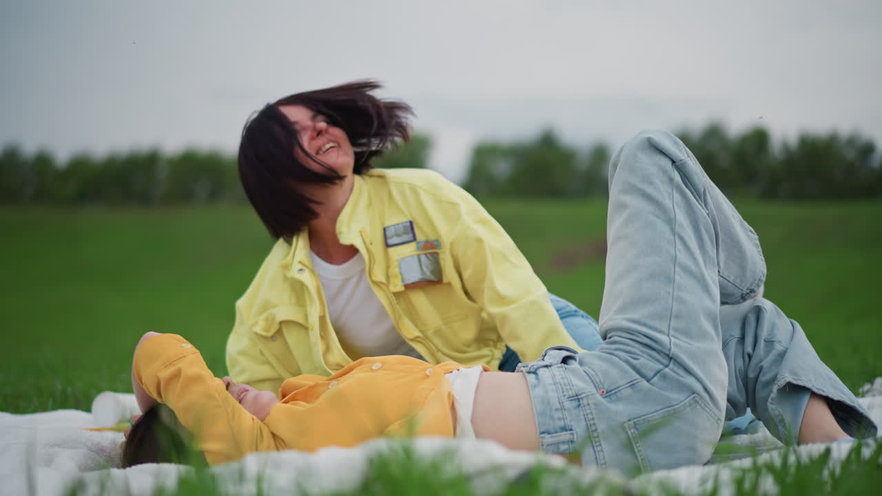 two women cuddling on blanket, tender kisses and embraces in open meadow with denim and yellow layers, overcast sky and green field, intimate romantic mood, candid closeup smiles, slow natural
