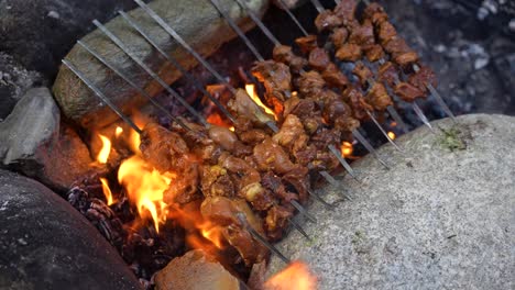 close-up of meat grilling over an open flame on a metal rack, surrounded by rocks, with flames and smoke