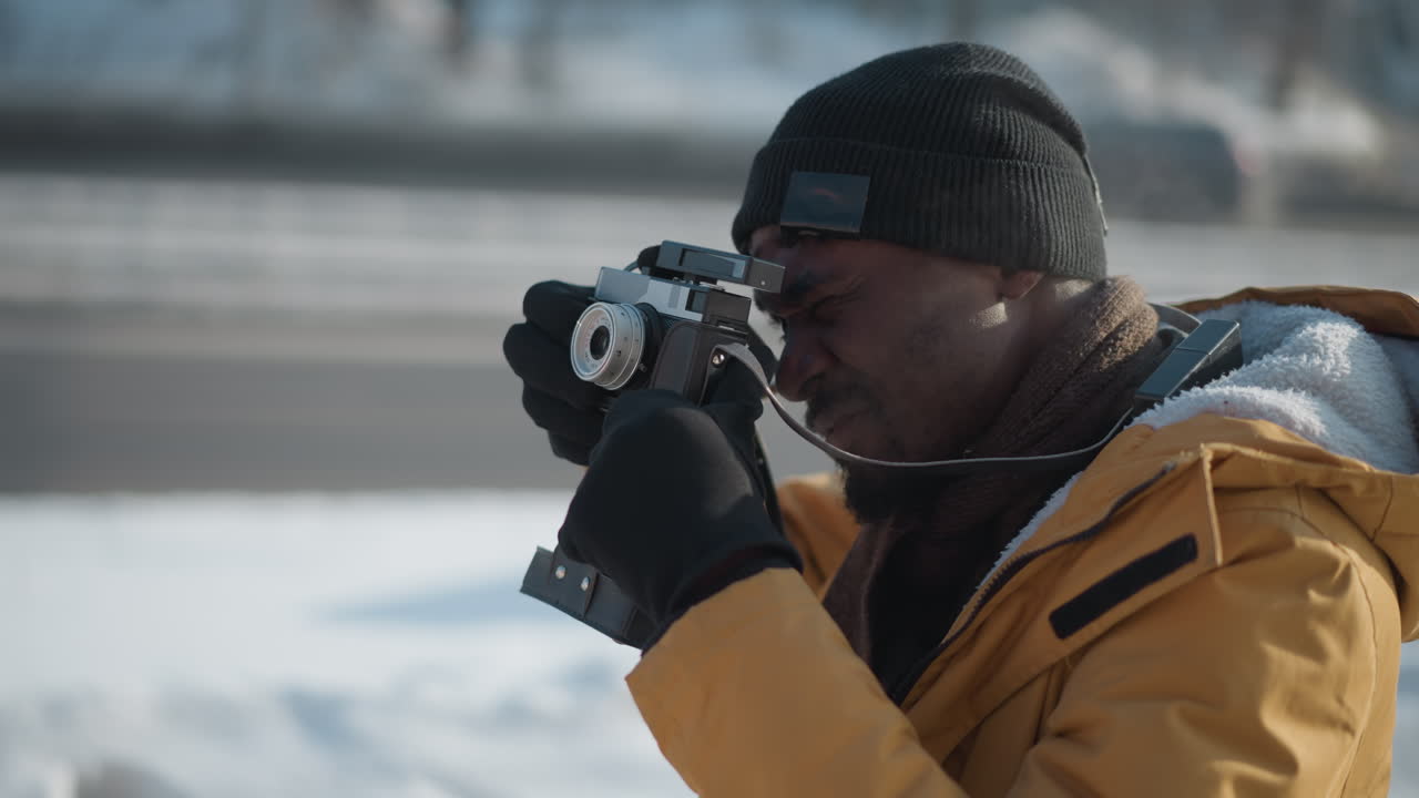 close up side view of operator holding camera adjusting lens with gloved hands walking along snow covered pavement beside busy street with blurred moving cars and frosty urban backdrop cold daylight