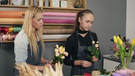 Atractiva-Mujer-Rubia-Con-Delantal-Parada-Con-Su-Compañera-De-Trabajo-En-El-Mostrador-Con-Una-Foto-Floral-Mientras-Arregla-Flores