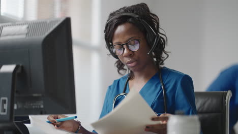 african american black female general practitioner in white coat sitting at desk in doctor's office and scrolling computer mouse while reading patient's medical history