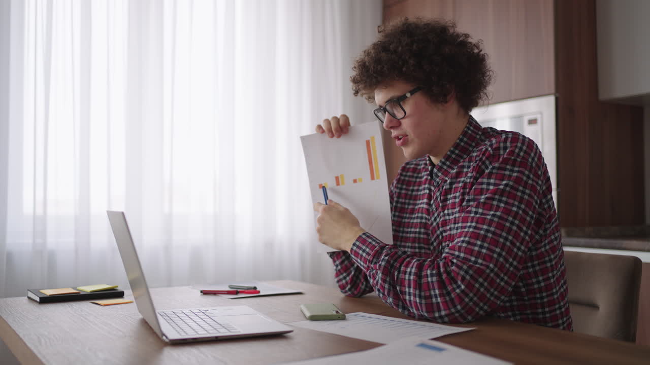 Curly - haired with glasses business man sitting at office from home desk looking at camera and pointing at a tablet with financial information displayed in graphical form column graph
