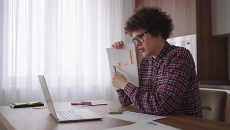 curly - haired with glasses business man sitting at office from home desk looking at camera and pointing at a tablet with financial information displayed in graphical form column graph