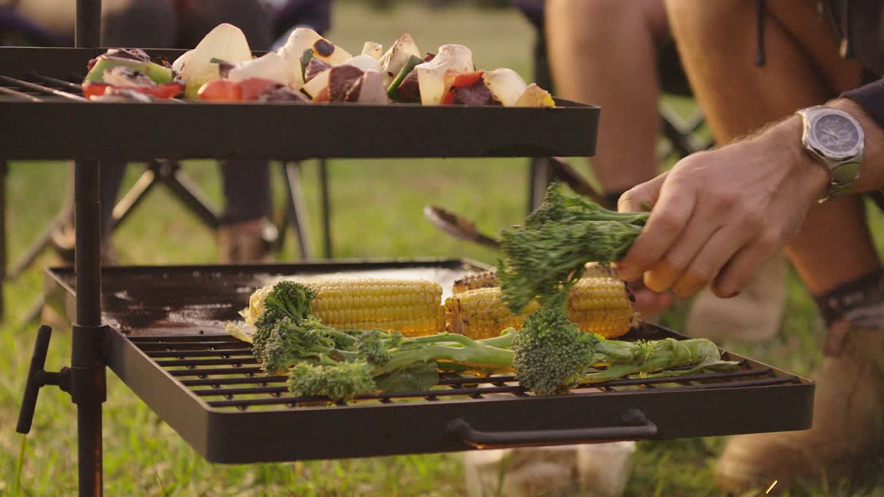 Placing broccolini vegetables on an open campfire BBQ to cook alongside some corn cobs and lamb kebab skewers