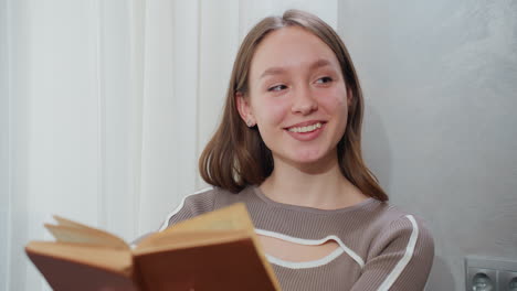 joven leyendo libro de portada marrón mira hacia arriba con una cálida sonrisa mientras está sentada cerca de la ventana con vidrio transparente, luz suave creando una atmósfera pacífica y acogedora en un interior elegante