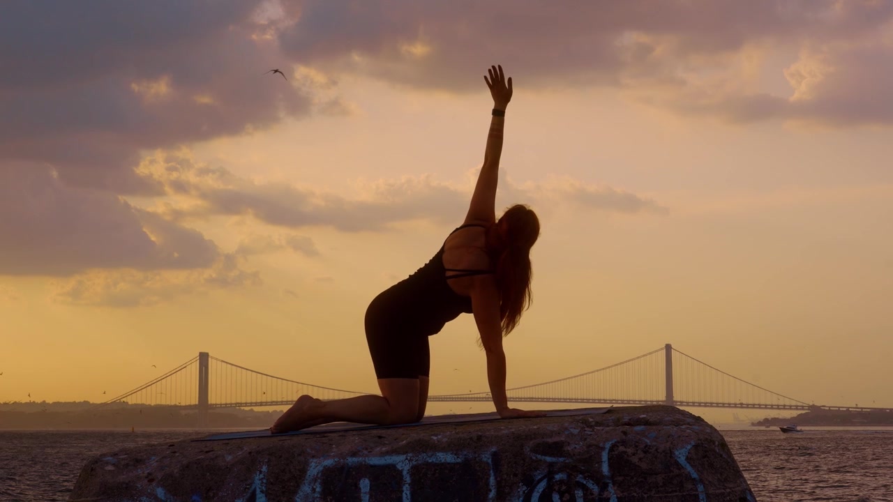 Donna che esegue la posizione yoga "infilare l'ago" al tramonto. Silhouette che pratica lo stretching delle spalle su un molo di pietra, Ponte di Verrazzano Narrows, cielo giallo, acqua calma.
