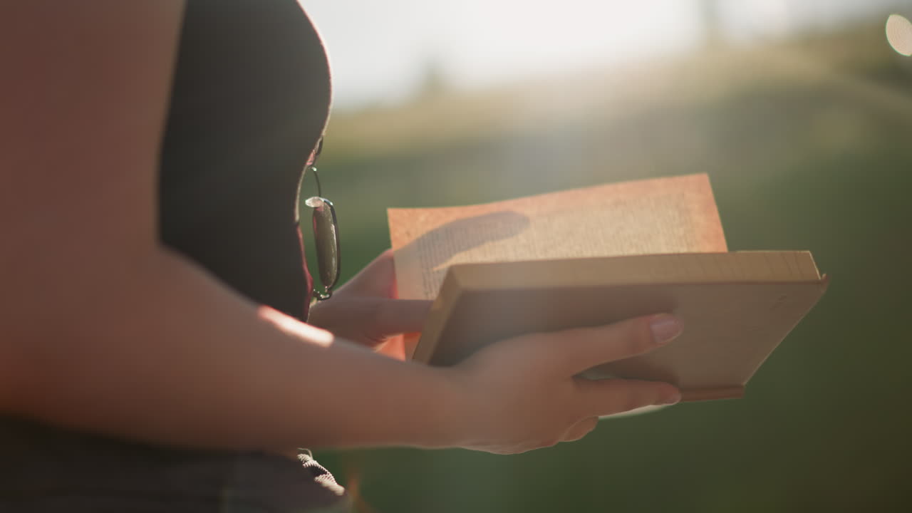 la luz del sol brilla desde la colina de verdor en un fondo borroso, reflejándose en el libro sostenido por una mujer con gafas colgando de su tapa negra mientras hojea las páginas del libro