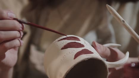 young woman potter painting on the ceramic cup with a brush and red color