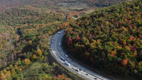 traffic rounds corner through mountains covered in autumn fall foliage, colorful leaves, trucks and cars on interstate turnpike highway road