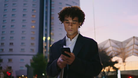 Apuesto-Hombre-De-Negocios-Deslizando-La-Pantalla-Del-Teléfono-Ciudad-Al-Atardecer.-Mensajes-De-Texto-Joven-Emprendedor