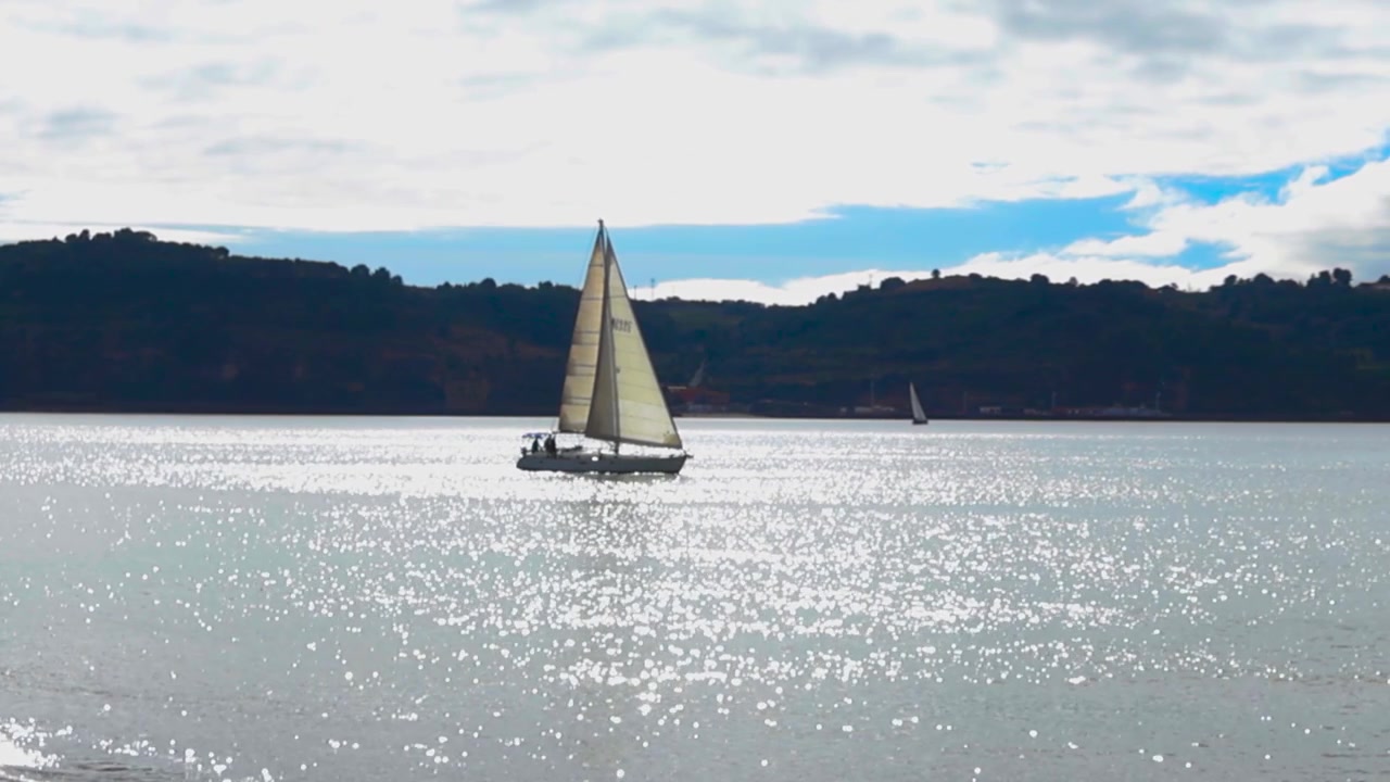 Vista panoramica di una barca a vela che scivola placidamente sulle acque scintillanti del fiume Tago a Lisbona, Portogallo, con il sole che si riflette sulla superficie e una costa in silhouette sullo sfondo