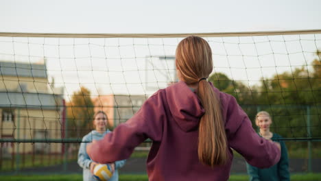 vista trasera de la dama en suéter marrón posando en la victoria en la cancha de voleibol al aire libre, el oponente mirando mientras uno sostiene el voleibol, mostrando el momento de triunfo y confianza