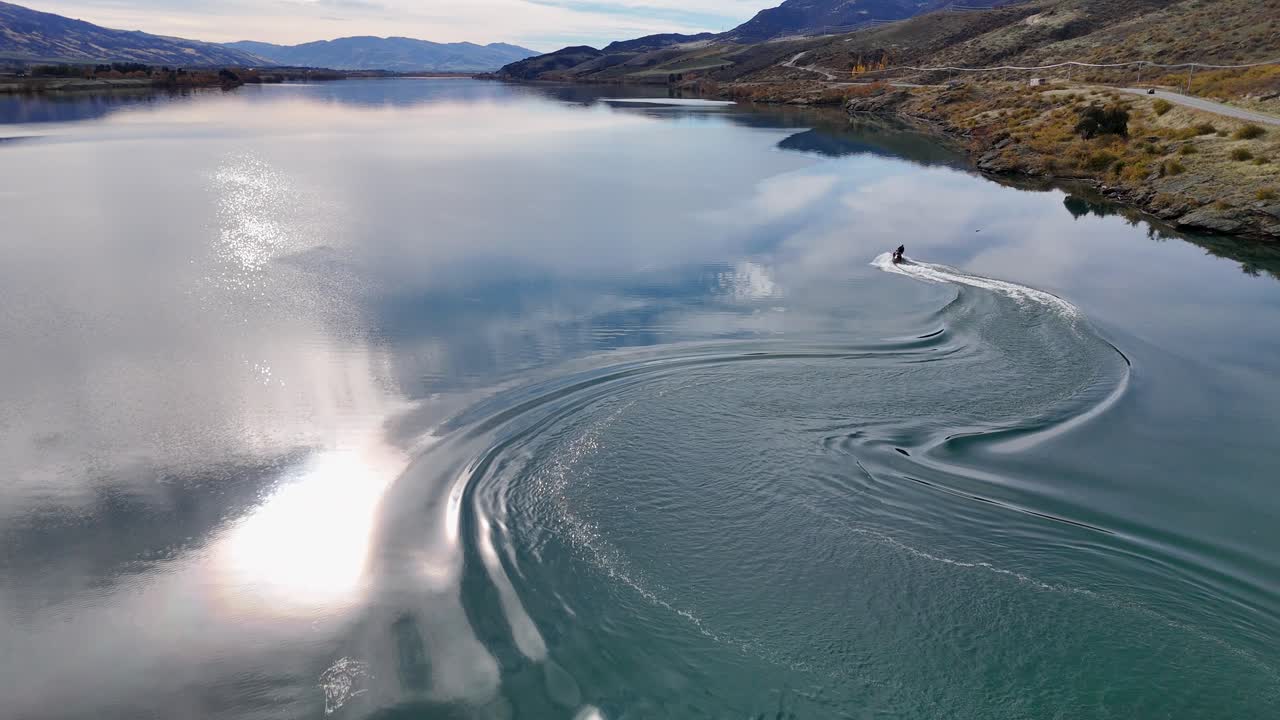 A jet ski glides across Lake Dunstan, creating ripples under bright sunlight, surrounded by serene autumn landscapes