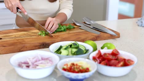 a person chops coriander on a wooden board surrounded by fresh vegetables in a bright kitchen