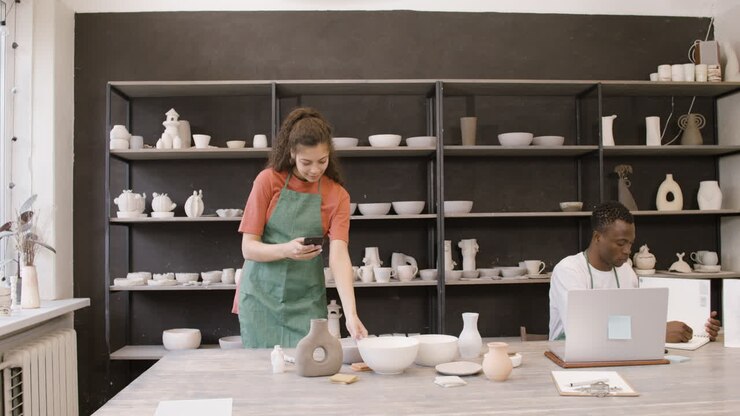 Woman Taking Photos Of Handmade Ceramic Pieces And Showing Pictures To Her Male Colleague Who Working On Laptop Computer In The Pottery Shop