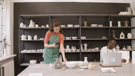 woman taking photos of handmade ceramic pieces and showing pictures to her male colleague who working on laptop computer in the pottery shop