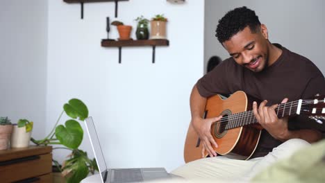 cheerful ethnic man playing guitar with laptop on sofa