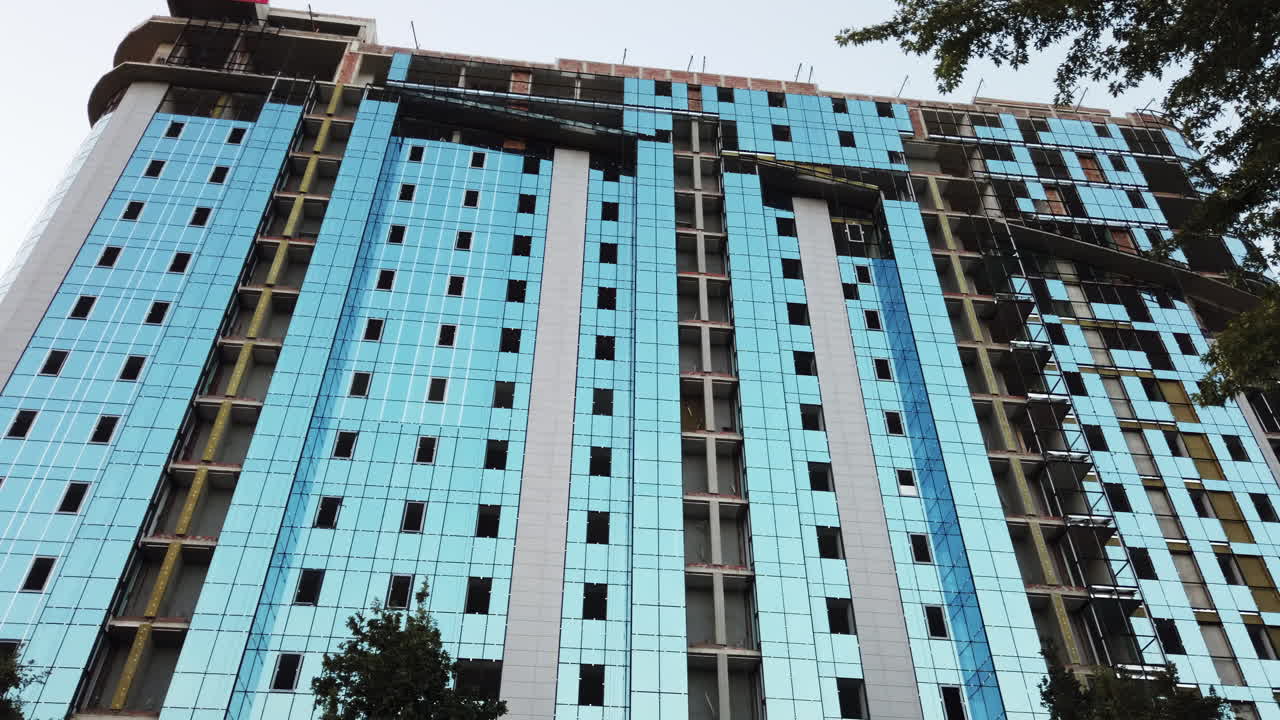 Workers are busy on the high-rise building, installing blue glass panels on the facade. The project is located in a bustling urban area and showcases modern architecture