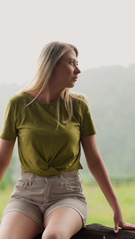 dreaming woman tourist in casual clothes enjoys wild nature sitting on log against lush field and mountain silhouette in rainy weather slow motion