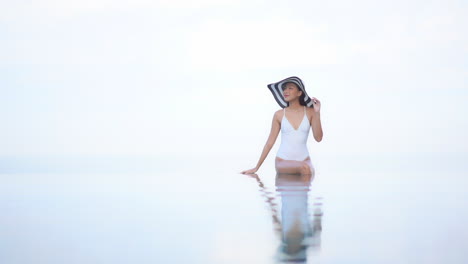 asian woman in a white bathing suit and floppy striped sunhat sits on the edge of the high rooftop infinity pool with feet in the water together with body reflection in water - copy space dream look