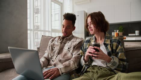 Happy young Black-skinned brunette man with stubble in a checkered cream shirt sits with his girlfriend and looks at something on his gray laptop while spending time together on the sofa in a modern studio apartment