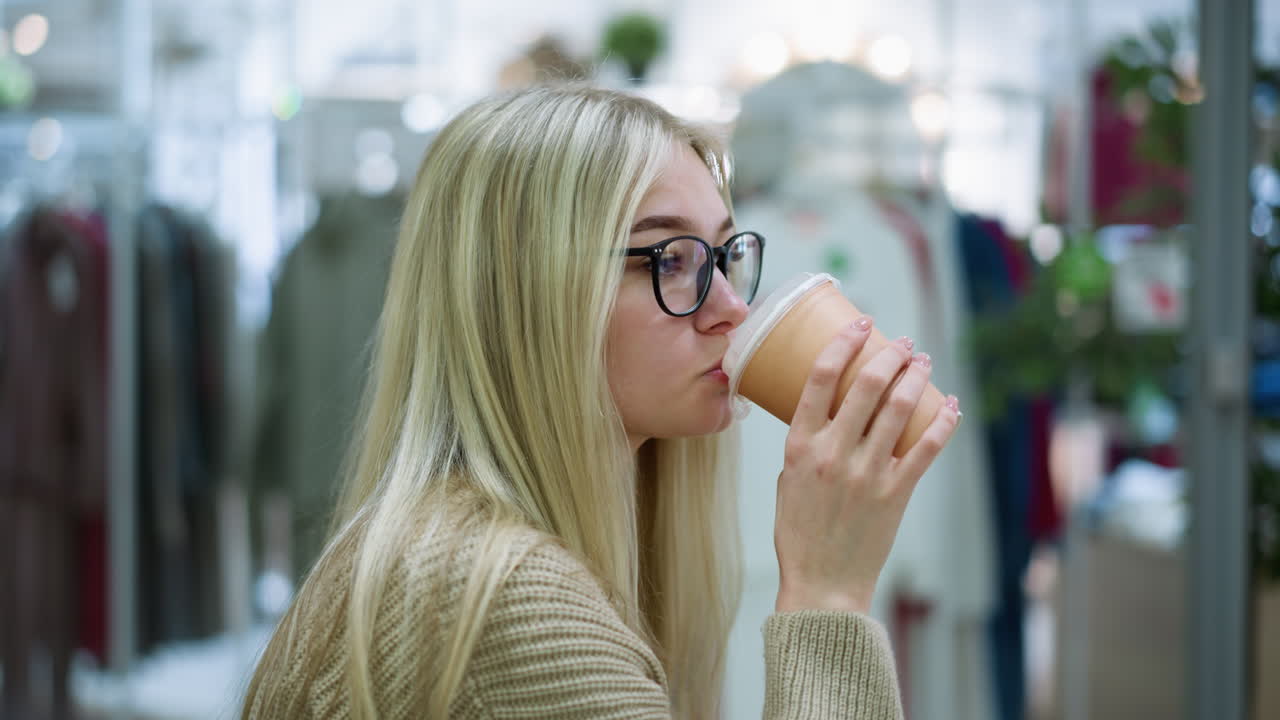 vista lateral de una joven con gafas bebiendo una bebida, mirando contemplativa en una tienda de ropa, con fondo de tienda bien iluminado y ropa expuesta en estantes
