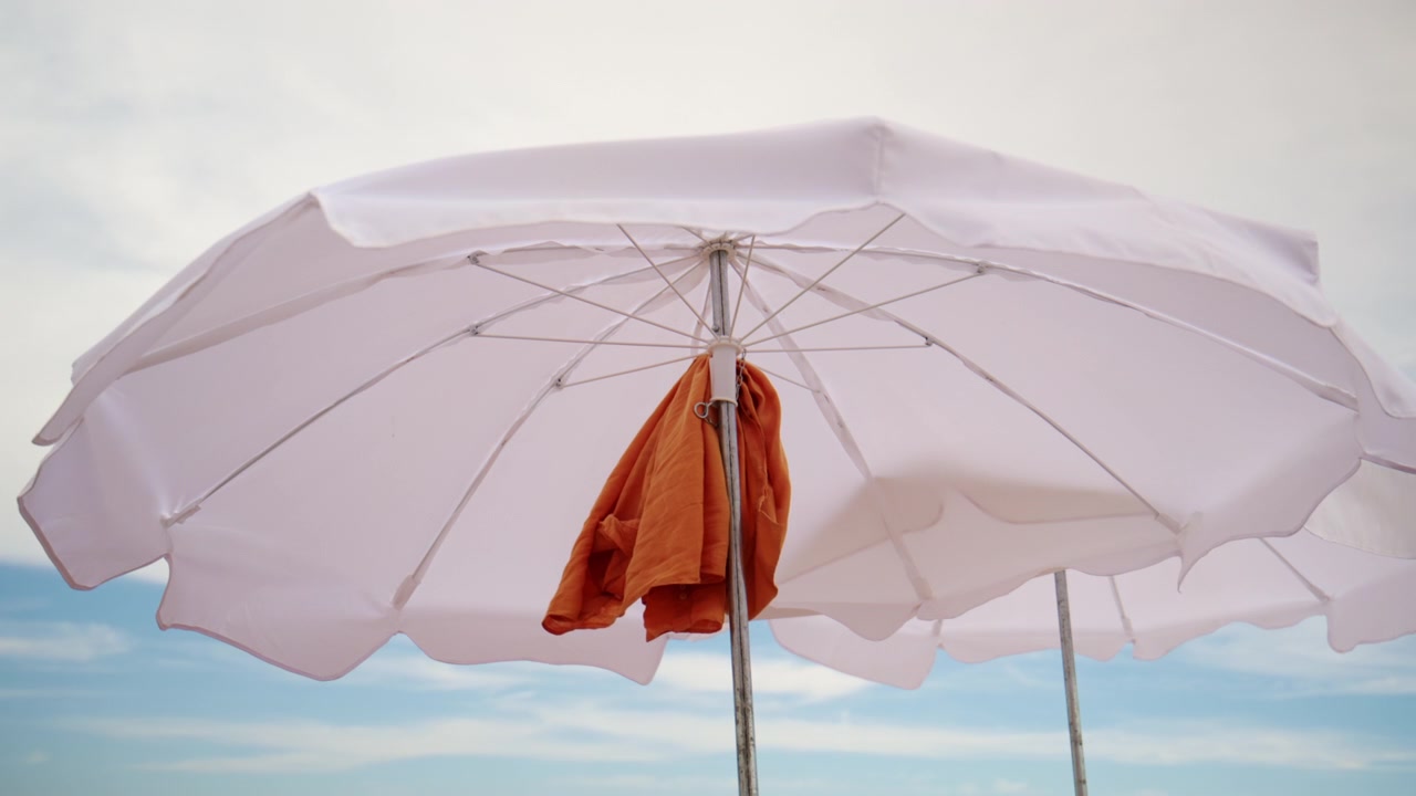 Ombrellone da spiaggia bianco con un asciugamano arancione appeso, che offre ombra contro un cielo blu, simboleggiando le vacanze estive e il relax