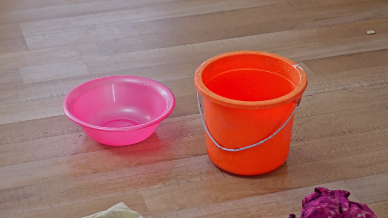 Orangener Eimer und pinke Wanne fangen Wasser von einem Deckenleck auf dem Holzboden auf