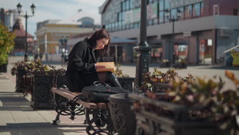 woman seated on bench in urban street reading a book and flipping pages, with her bag resting beside her, white leather blowing in the wind beneath her chair, peaceful street setting