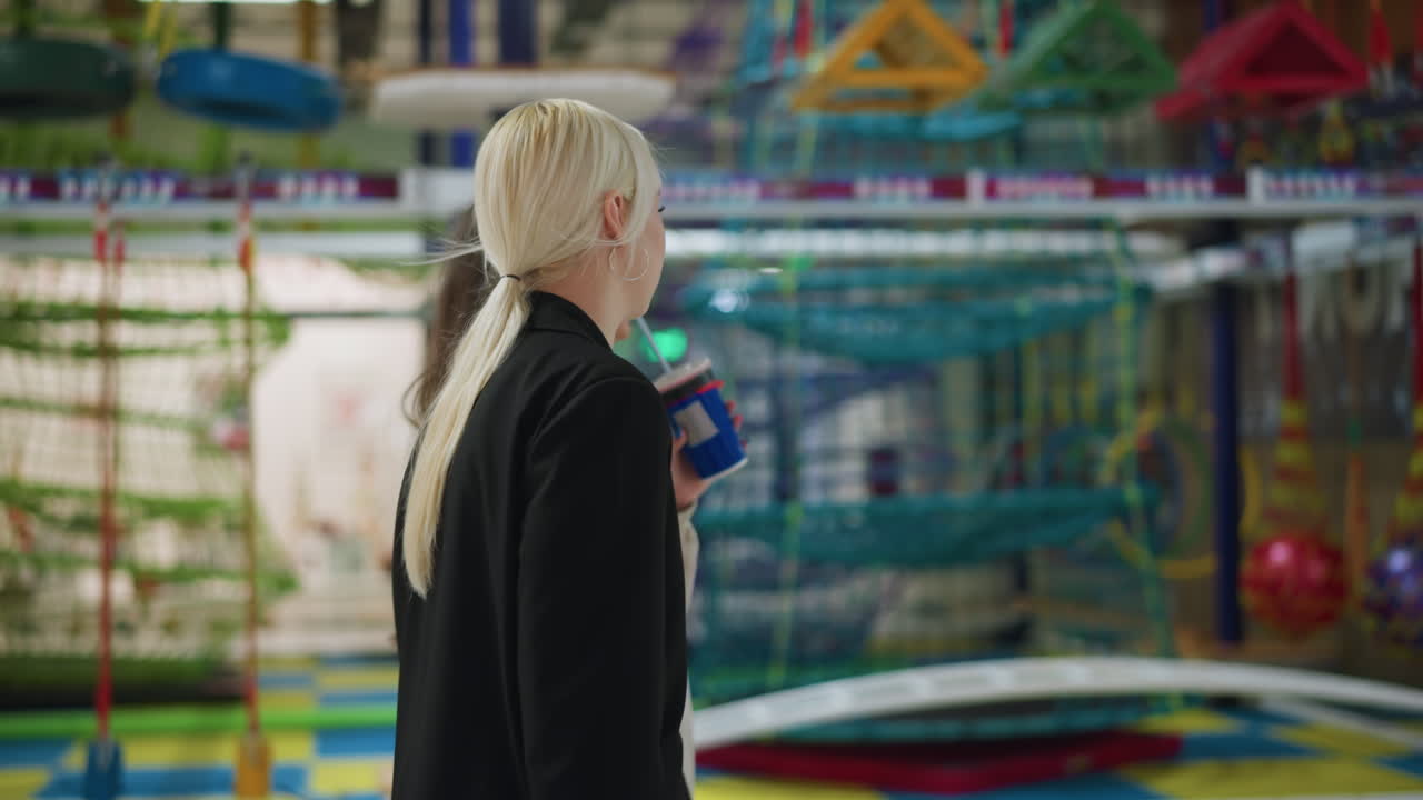 Side view of siblings walking together through playground while swinging hands, one holding cola drink, sharing relaxed bond in vibrant environment filled with hanging ropes and play structures