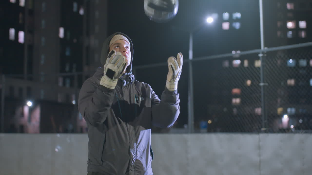 futbolista joven con guantes lanza una pelota de fútbol al aire durante la práctica nocturna en un campo urbano, el fondo incluye edificios altos, cercas de cadena y iluminación bokeh suave