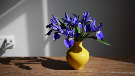 a bouquet of purple irises in a yellow vase on a wooden table with sunlight and shadows