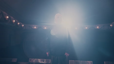 aged actor performing in theater pan right view of senior man gesticulating and talking while standing on stage in spotlight during dramatic performance in theater