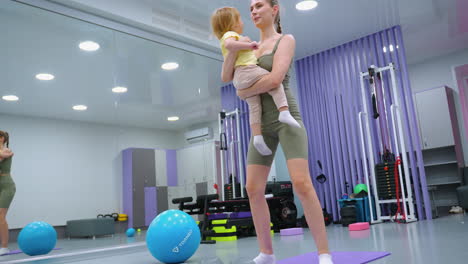la madre realiza sentadillas mientras sostiene a la hija, participando en una rutina de ejercicio, el niño se vuelve hacia la madre mientras se mueve arriba y abajo, equipos de fitness, espejos de gimnasio y luces brillantes