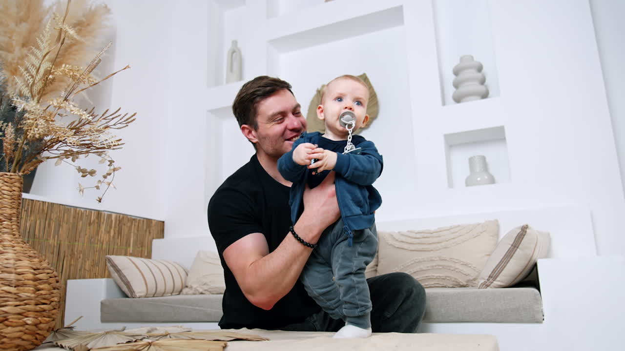 Caucasian male holds an infant child standing on the coffee table. Cute kid with pacifier smiles adorably. Low angle view.