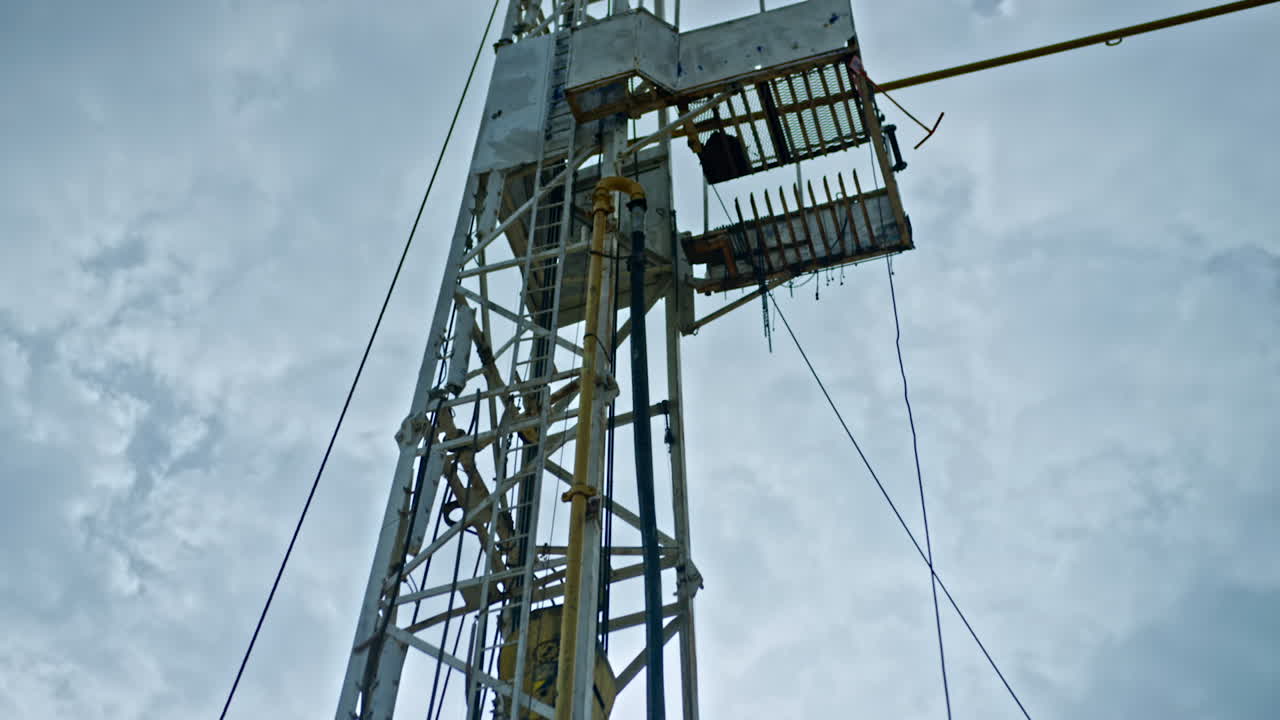 Metal tower with pipes and wires attached. Low angle view on the oil rig at the backdrop of cloudy sky.