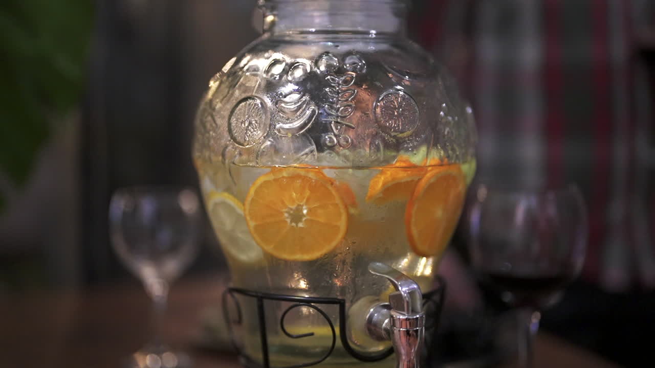 Right panning closeup clip of refreshing orange infused beverage stored in large glass dispenser, with blurred background at restaurant.