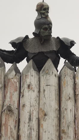 man in armor holds spear and surveys area behind high log fence standing near burnt roof rain. dark knight attacks inhabitants of medieval village low angle shot