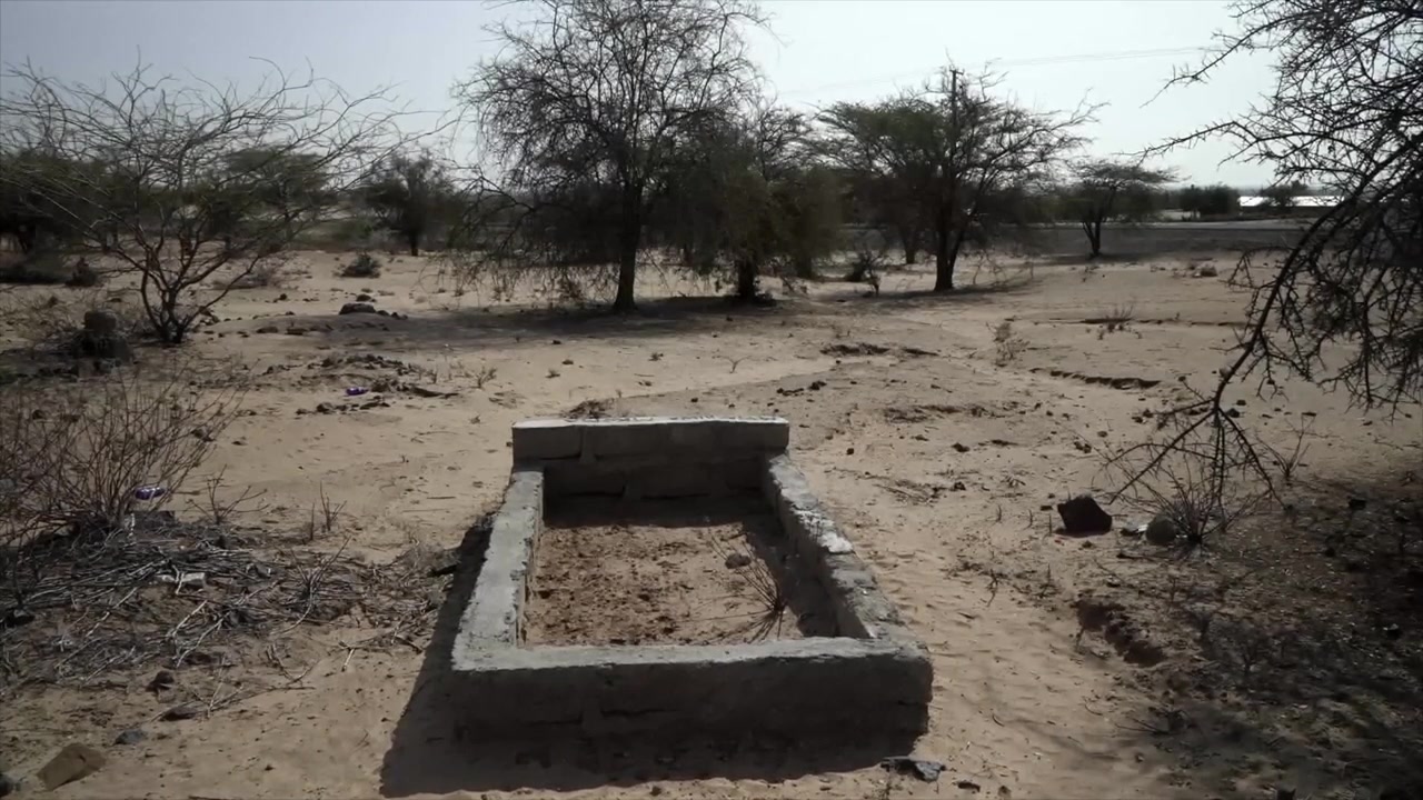 Sebuah makam atau kuburan beton sederhana terletak di lanskap kering berpasir Gurun Turkana, Kenya, dikelilingi oleh pohon akasia berduri yang jarang di bawah langit berkabut.