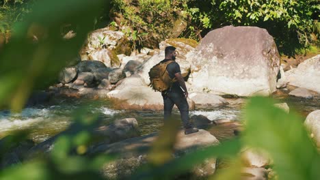 slow motion young male hiker along mountain river surounded by boulders and trees, looking around during hike in jungle