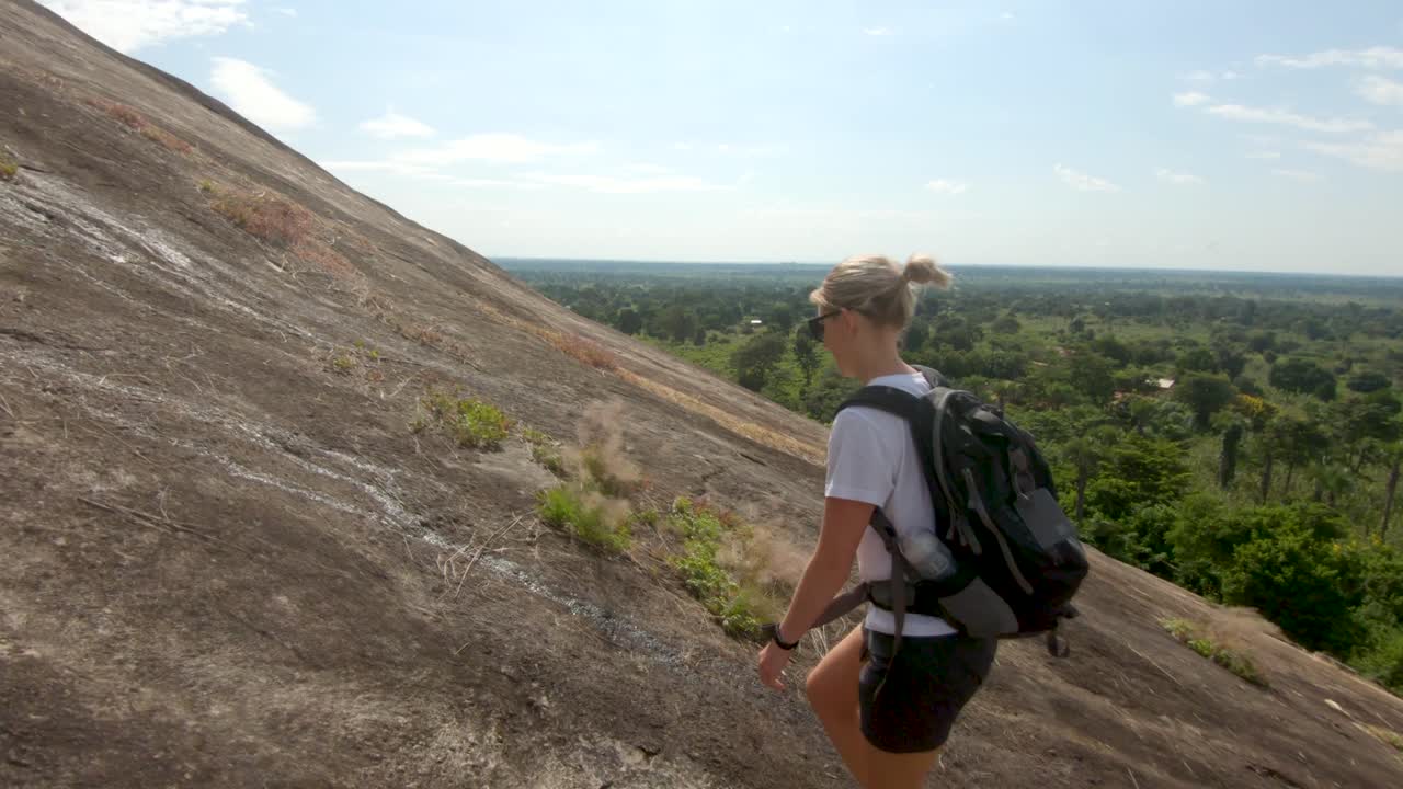 toma amplia siguiendo a una turista occidental rubia mientras sube una gran roca de granito en las zonas rurales de áfrica oriental