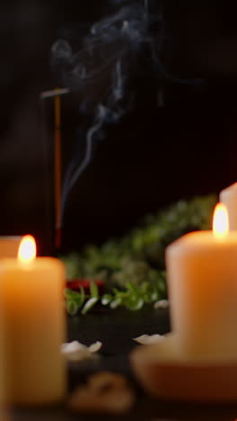 Vertical-Video-Shot-Of-Person-Picking-Up-Towel-With-Lit-Candles-With-Scattered-Petals-Incense-Stick-Against-Dark-Background-As-Part-Of-Relaxing-Spa-Day-Decor