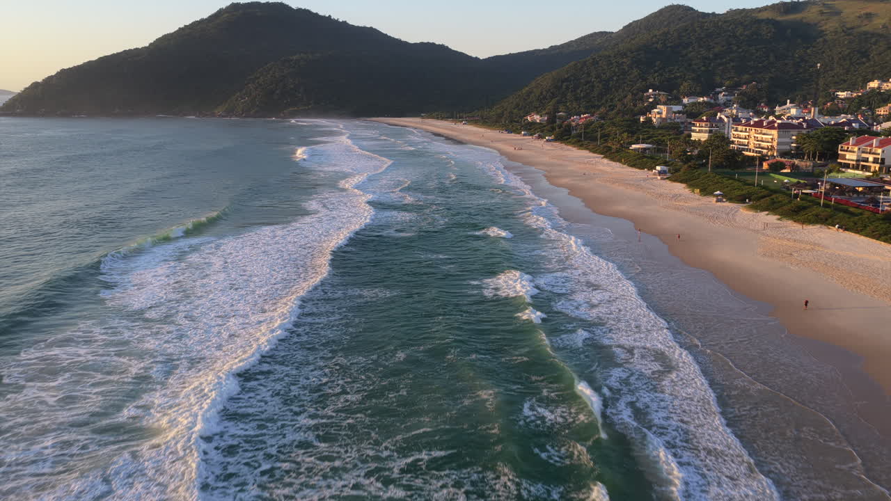 Praia Brava di Florianópolis, Santa Catarina, Brasil di Samudra Atlantik dengan ombak bergulir dan garis pantai yang membentang di bawah pegunungan Serra do Mar dalam cahaya matahari terbit yang hangat, penerbangan drone maju